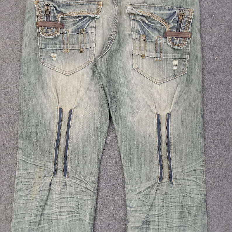 May include: Light blue denim jeans with distressed details, featuring decorative zippers on the front and pockets with zipper and leather accents. The jeans have a faded wash and a classic five-pocket design.