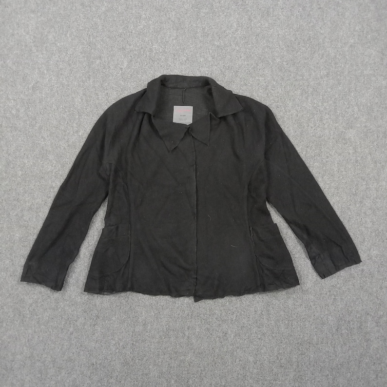 May include: A solid black long-sleeved jacket featuring a pointed collar, a front button closure, and two side pockets. The garment has a structured fit with a straight hemline and a small white brand label attached to the inside of the collar.