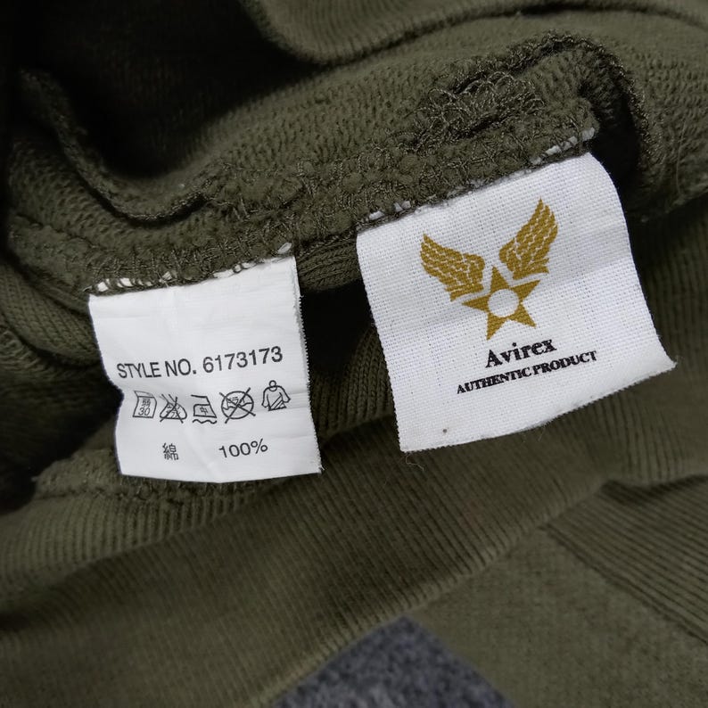 May include: Close-up of a green garment with two attached tags. One tag displays the text "STYLE NO. 6173173" along with care symbols and "綿 100%." The other tag features a gold emblem with wings and a star, and the text "Avirex AUTHENTIC PRODUCT."