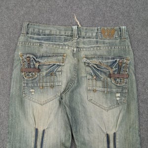 May include: Faded blue denim jeans with multiple pockets, zippers, and decorative stitching. The back pockets have a unique design with zippers and brown leather accents. The jeans feature a distressed look with slight fading and wear.