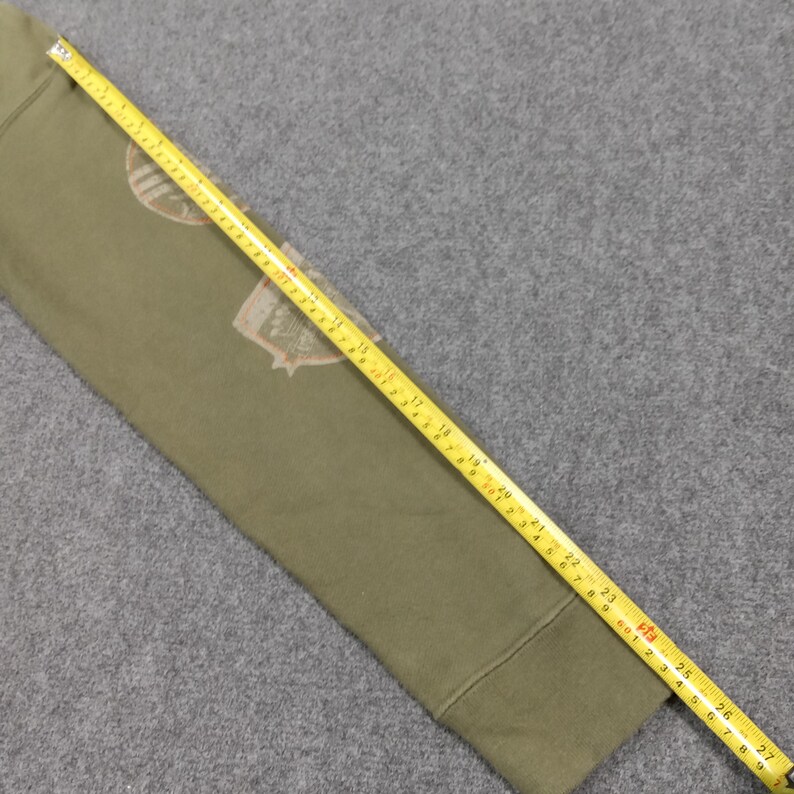 May include: Olive green sweatshirt sleeve with a beige and brown graphic design. A yellow measuring tape is laid across the sleeve, showing measurements in inches. The sleeve is on a gray surface.