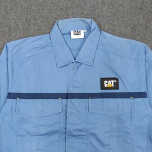 Puede incluir: Camisa de trabajo azul claro con dos bolsillos en el pecho y una franja azul marino en el pecho. La camisa presenta el logotipo "CAT" en el pecho y el cuello. Tiene una botonadura y un cuello.