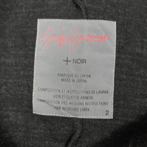 May include: A rectangular light gray clothing label sewn onto a dark charcoal gray fabric. The label features a red signature logo at the top, the text + NOIR, and the phrases FABRIQUE AU JAPON, MADE IN JAPAN, and instructions to see the attached label.
