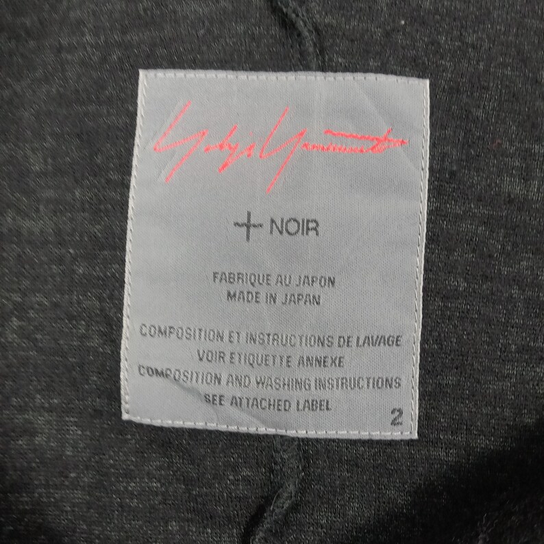 May include: A rectangular light gray clothing label sewn onto a dark charcoal gray fabric. The label features a red signature logo at the top, the text + NOIR, and the phrases FABRIQUE AU JAPON, MADE IN JAPAN, and instructions to see the attached label.