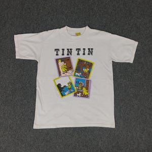 May include: White t-shirt featuring the text "TINTIN" above four colorful square illustrations. The illustrations depict scenes with characters and a dog. The shirt has a crew neck and short sleeves.