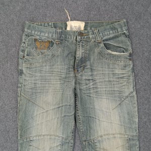 May include: Light blue denim jeans with a distressed wash. Features include a decorative embroidered pocket detail and a small tag. The jeans have a button closure and a classic five-pocket design.