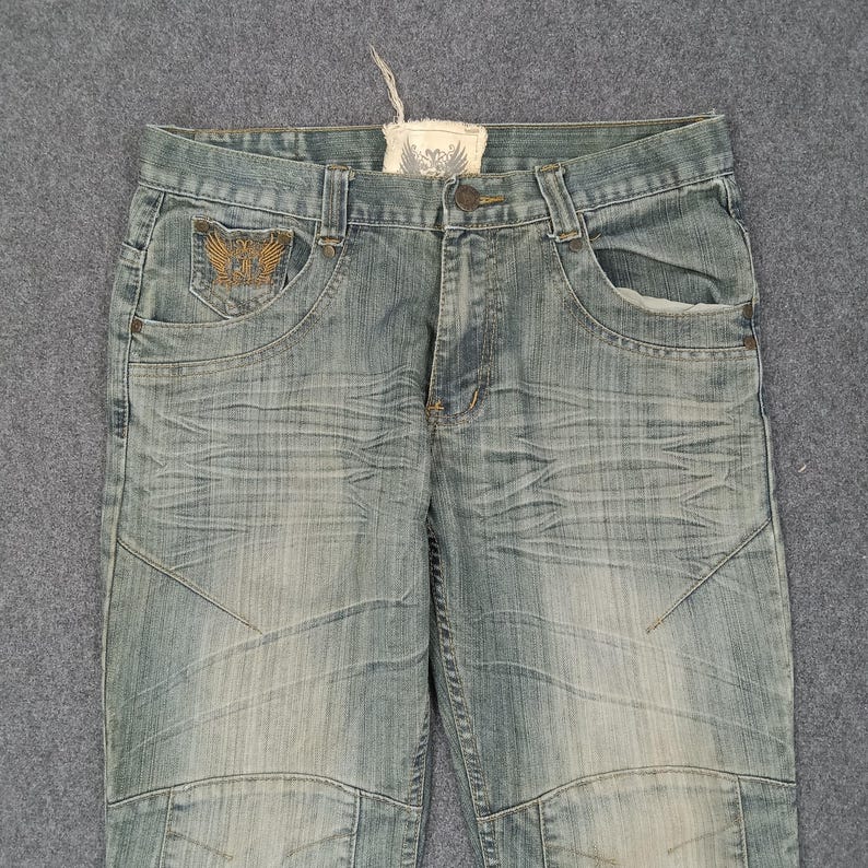 May include: Light blue denim jeans with a distressed wash. Features include a decorative embroidered pocket detail and a small tag. The jeans have a button closure and a classic five-pocket design.