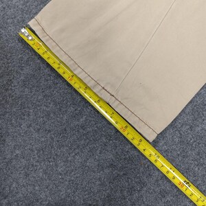 May include: Close-up of the bottom edge of a pair of beige pants with a red stitch detail. A yellow measuring tape is laid across the fabric, showing measurements in inches.