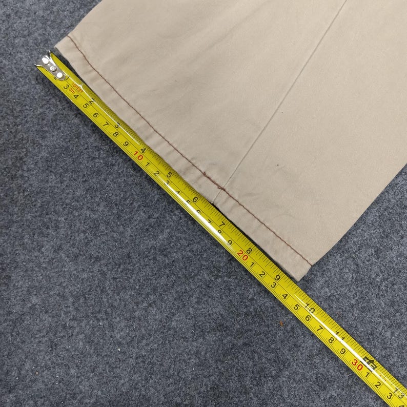 May include: Close-up of the bottom edge of a pair of beige pants with a red stitch detail. A yellow measuring tape is laid across the fabric, showing measurements in inches.