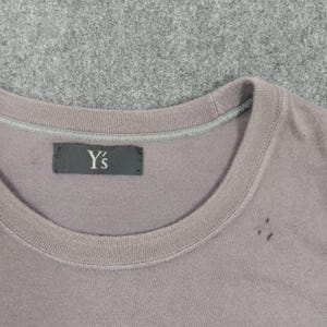 Puede incluir: Primer plano de una camiseta gris con cuello redondo. La camiseta tiene una peque&ntilde;a etiqueta negra con el texto blanco "Y's". La camiseta est&aacute; hecha de un material suave y tiene unos peque&ntilde;os agujeros cerca del cuello.