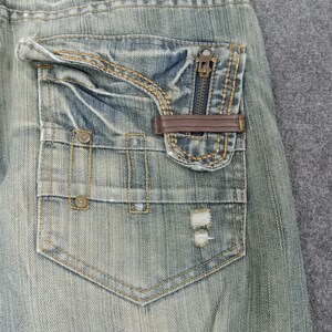 May include: Close-up of a light blue denim jeans pocket. The pocket features a zipper closure, brown leather accent, and decorative stitching. The denim has a distressed, worn appearance with a small tear near the bottom.