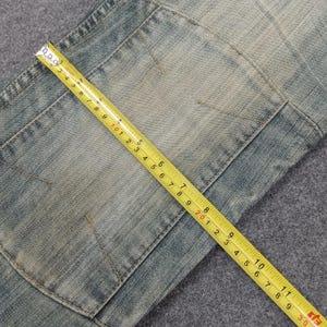 May include: Close-up of light blue denim jeans with a patch pocket. A yellow measuring tape is laid across the jeans, showing measurements in inches. The jeans have a faded, worn appearance.
