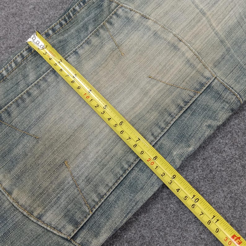 May include: Close-up of light blue denim jeans with a patch pocket. A yellow measuring tape is laid across the jeans, showing measurements in inches. The jeans have a faded, worn appearance.
