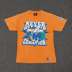 Peut inclure: T-shirt orange avec un graphisme d'haltérophile et l'inscription "NEVER GIVE UP CENATION". Le t-shirt porte un petit logo WWE sur le col et un écusson sur la manche.