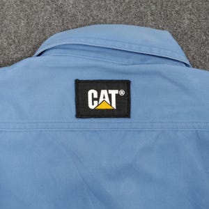 Puede incluir: Una camisa de trabajo azul claro con cuello y un parche negro con el logotipo de CAT en blanco y amarillo. La camisa tiene una costura cosida en la espalda.