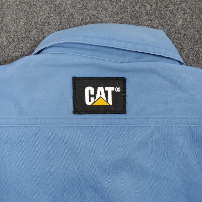 Puede incluir: Una camisa de trabajo azul claro con cuello y un parche negro con el logotipo de CAT en blanco y amarillo. La camisa tiene una costura cosida en la espalda.