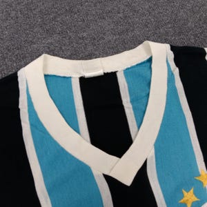 May include: A striped v-neck shirt with blue, white, and black stripes. The shirt has a white collar and two gold stars on the right side.