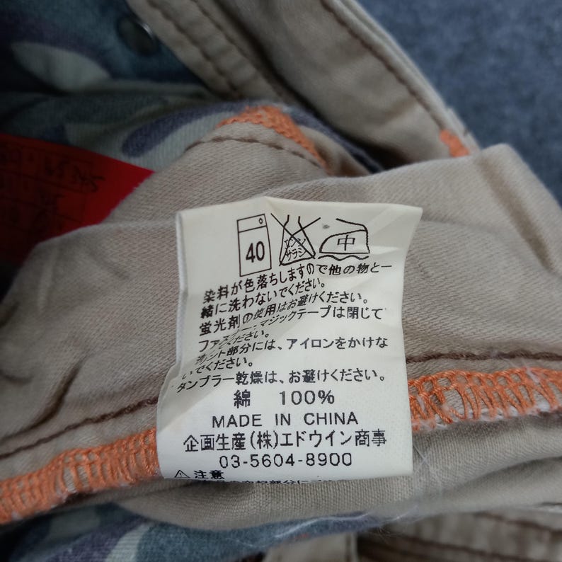 May include: A beige clothing care label with black and red text. The label has instructions for washing, drying, and ironing. The label also includes the size 40, the fabric content (100% cotton), and the manufacturer's information: "Made in China,  企画生産(株)エドウイン商事 03-5604-8900".