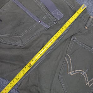 Op de afbeelding: Een donkergroene cargo broek met een meetlint eroverheen gelegd. De broek heeft meerdere zakken en een contrasterend grijs stoffen detail op de voorzak.
