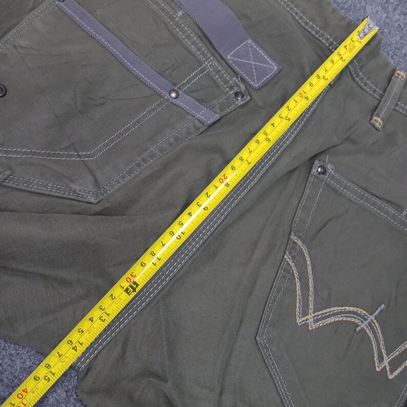 Op de afbeelding: Een donkergroene cargo broek met een meetlint eroverheen gelegd. De broek heeft meerdere zakken en een contrasterend grijs stoffen detail op de voorzak.