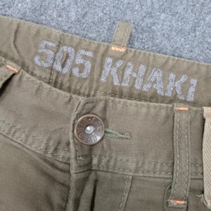 May include: Close-up of a pair of khaki pants with a button and a label that reads "505 KHAKI".