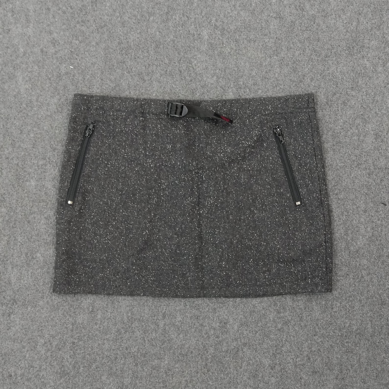 Pode incluir: Minissaia cinza com dois bolsos com z&iacute;per e fecho de fivela preto. A saia tem uma textura salpicada e um design simples e funcional, adequado para atividades ao ar livre.
