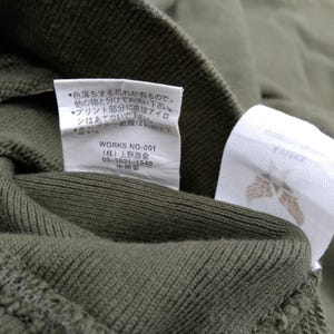 May include: Close-up of olive green fabric with two white tags attached. One tag has Japanese text and the other has a brown floral design. The fabric appears to be a knit material. The Japanese text includes washing instructions.