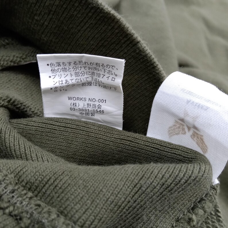 May include: Close-up of olive green fabric with two white tags attached. One tag has Japanese text and the other has a brown floral design. The fabric appears to be a knit material. The Japanese text includes washing instructions.