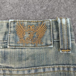 May include: Close-up of denim jeans with a distressed wash. The jeans feature a brown leather patch with a winged emblem and decorative stitching. The jeans have a classic five-pocket design and belt loops.