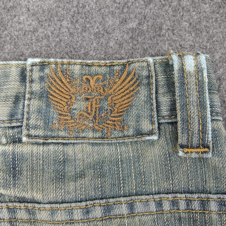 May include: Close-up of denim jeans with a distressed wash. The jeans feature a brown leather patch with a winged emblem and decorative stitching. The jeans have a classic five-pocket design and belt loops.