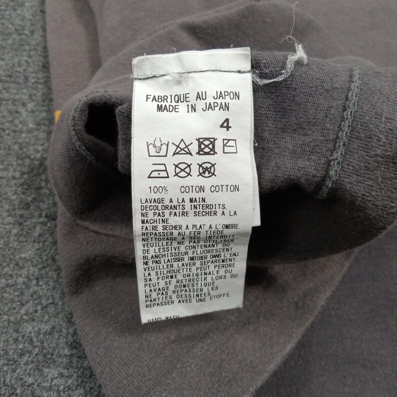 Puede incluir: Primer plano de una etiqueta de prenda gris con texto en franc&eacute;s e ingl&eacute;s. La etiqueta dice "FABRIQUE AU JAPON MADE IN JAPAN" e incluye instrucciones de lavado. La etiqueta est&aacute; adherida a una tela gris.