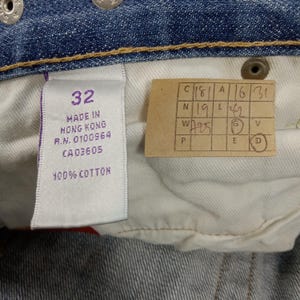 Op de afbeelding: Close-up van de binnenkant van een blauwe spijkerbroek. Een wit label met het nummer "32" en de tekst "MADE IN HONG KONG" is zichtbaar. Een klein, rechthoekig label met handgeschreven cijfers en letters is ook aanwezig. De jeans is gemaakt van 100% katoen.
