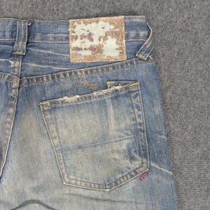 Op de afbeelding: Close-up van een lichtblauwe denim jeans met een distressed look. Voorzien van een achterzak en een versleten leren patch. De jeans vertoont tekenen van slijtage, met vervaagde plekken en gerafelde randen, wat hem een vintage uitstraling geeft.
