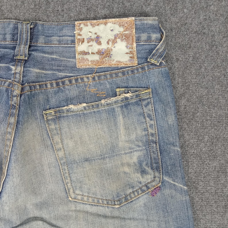 Op de afbeelding: Close-up van een lichtblauwe denim jeans met een distressed look. Voorzien van een achterzak en een versleten leren patch. De jeans vertoont tekenen van slijtage, met vervaagde plekken en gerafelde randen, wat hem een vintage uitstraling geeft.