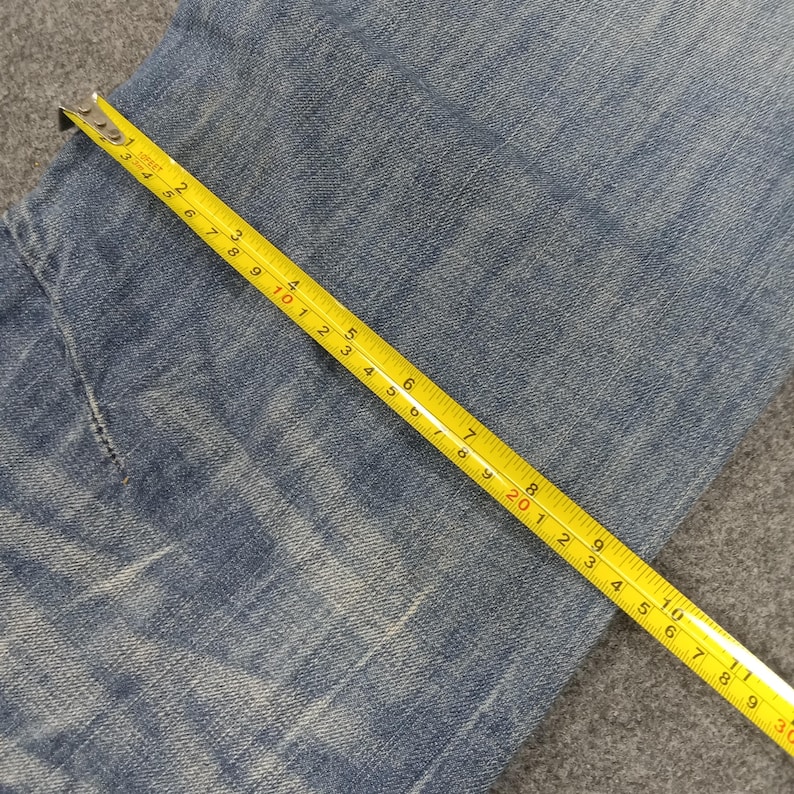 Op de afbeelding: Close-up van een lichtblauwe spijkerbroek met een gele meetlint over de stof. De tape toont metingen in centimeters, met de cijfers 1 tot en met 25 duidelijk zichtbaar. De jeans lijkt versleten.