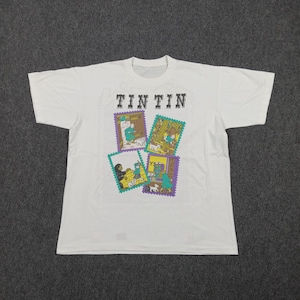 May include: White t-shirt featuring the word "TINTIN" above four colorful postage stamp-style illustrations. The illustrations depict scenes with characters and a dog. The shirt is made of a soft material.