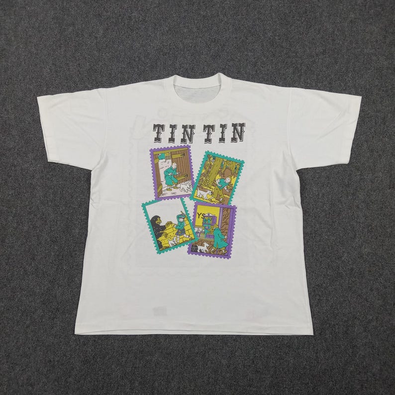 May include: White t-shirt featuring the word "TINTIN" above four colorful postage stamp-style illustrations. The illustrations depict scenes with characters and a dog. The shirt is made of a soft material.