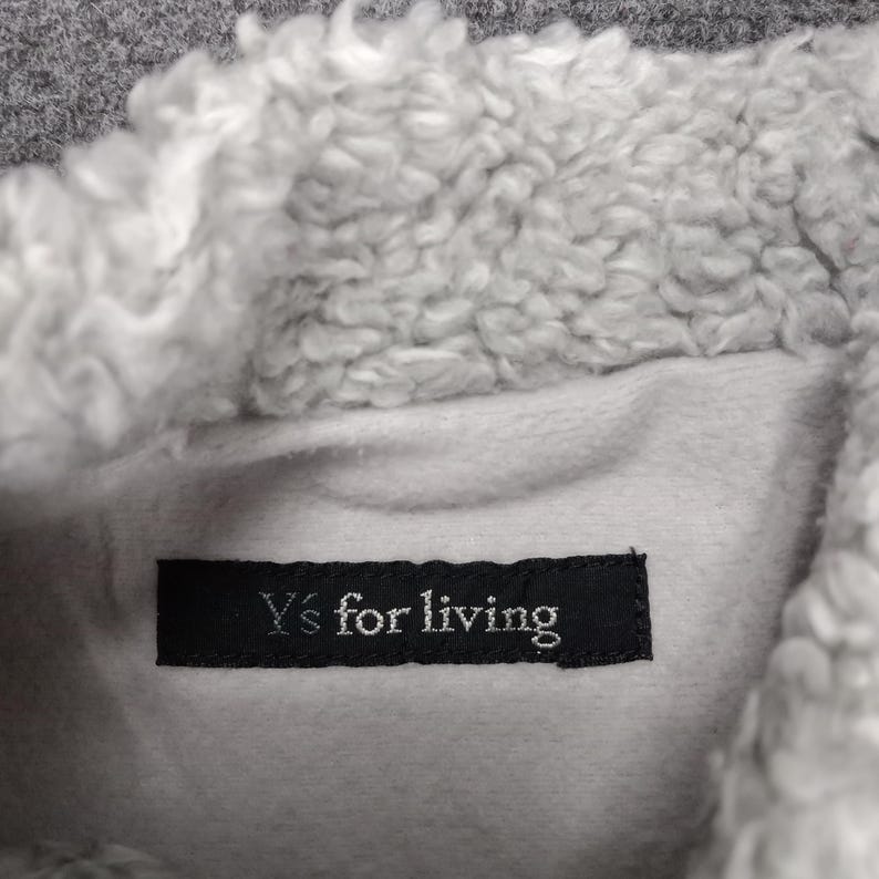 Op de afbeelding: Een close-up van een grijze fleecejas met een witte sherpa voering. Het label luidt "Y's for living".