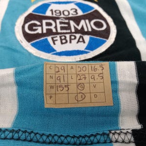 May include: A blue and white striped jersey with a black and white stripe. The jersey has a large circular patch with the text "1903 GR&Ecirc;MIO FBPA". The jersey also has a size tag with the text "C 29 A 20 16.5 N 91 L 27 9.5 W 155 G V E D".