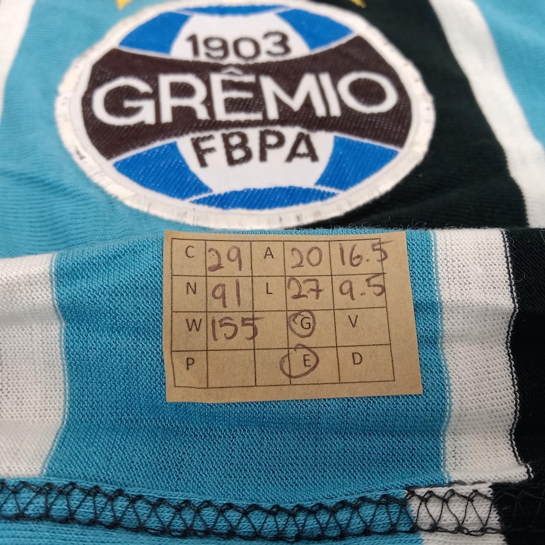 May include: A blue and white striped jersey with a black and white stripe. The jersey has a large circular patch with the text "1903 GR&Ecirc;MIO FBPA". The jersey also has a size tag with the text "C 29 A 20 16.5 N 91 L 27 9.5 W 155 G V E D".