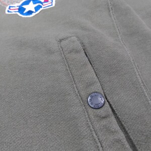 May include: Close-up of an olive green jacket with a patch featuring a star and wings. The jacket has a pocket and a button with the text "U.S. AIR FORCE". The fabric appears to be a soft, textured material.