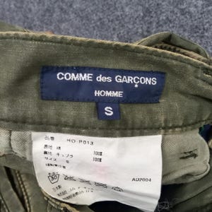 May include: Green cargo trousers with a black label that reads "COMME des GAR&Ccedil;ONS HOMME*" and a size tag that reads "S". The trousers have a white tag with the text "HO-P013", "100% cotton", "100% cupro", "S", and "AD2004".