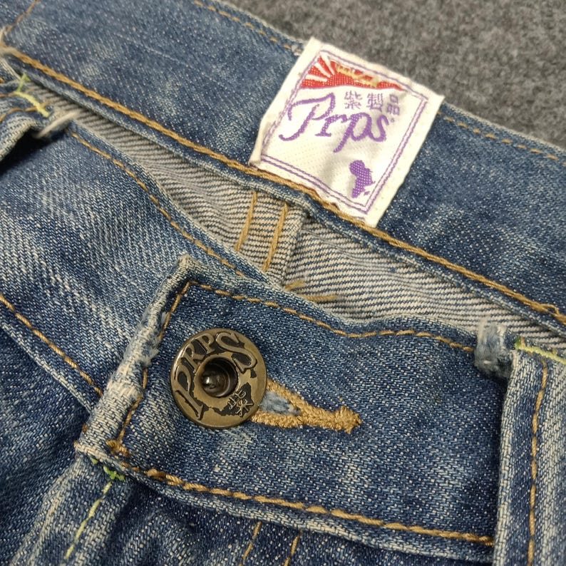 Op de afbeelding: Close-up van een blauwe spijkerbroek met een knoop en een label. De knoop is bronskleurig met een reli&euml;fdesign. Het label heeft het woord "Prps" in paarse letters en Japanse tekst.