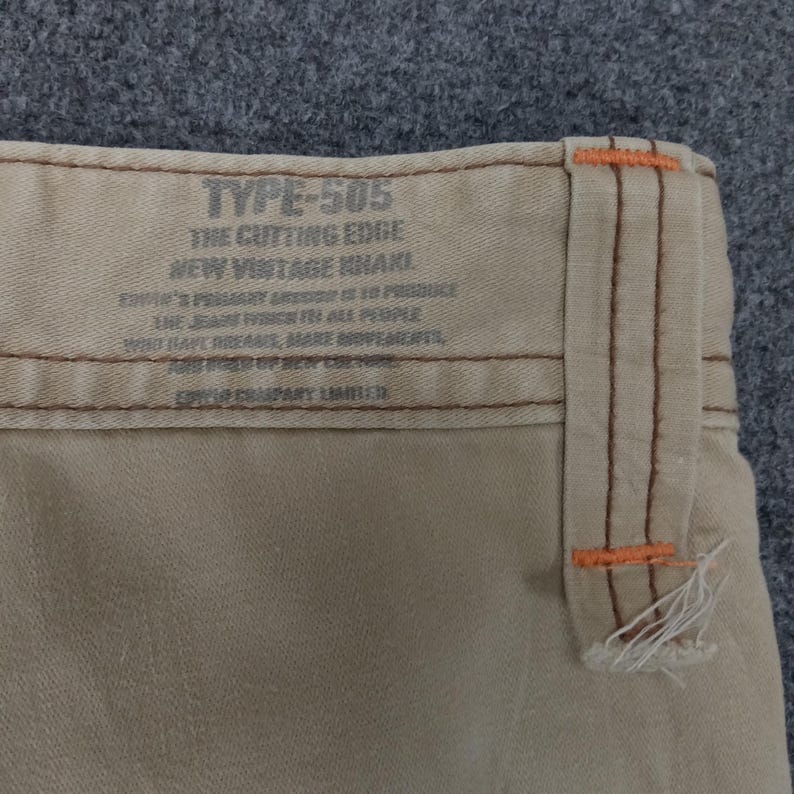 May include: Close-up of a beige pair of pants with a brown and orange stitched seam. The label reads "TYPE-505 THE CUTTING EDGE NEW VINTAGE KHAKI" and includes additional text about the product.