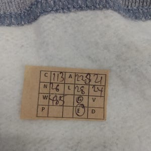 May include: Close-up of a light gray fabric with a blue trim. A small, rectangular tag is attached, displaying handwritten numbers and letters in black ink. The tag appears to be a size or measurement label.