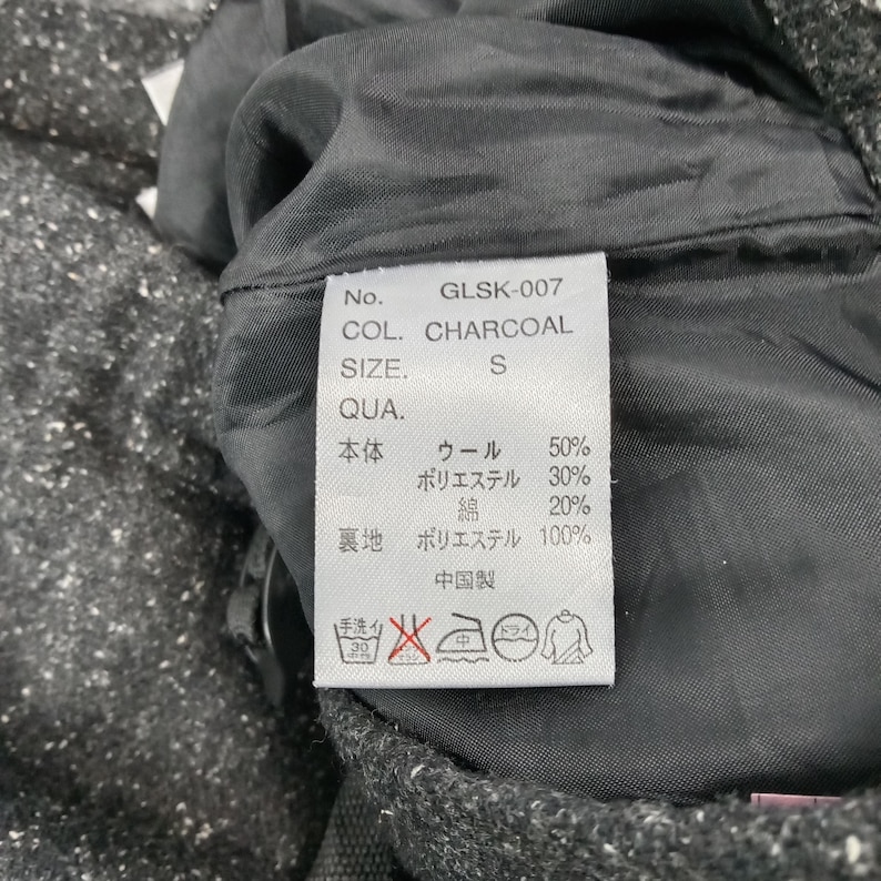 Pode incluir: Close-up de uma etiqueta de vestu&aacute;rio com o texto "GLSK-007 COL. CHARCOAL SIZE. S". A etiqueta lista a composi&ccedil;&atilde;o do tecido: l&atilde;, poli&eacute;ster e algod&atilde;o. As instru&ccedil;&otilde;es de lavagem tamb&eacute;m s&atilde;o vis&iacute;veis.