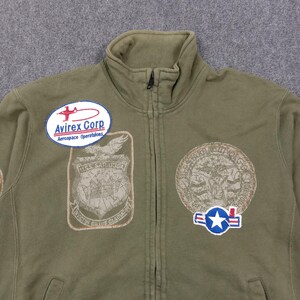 May include: Olive green zip-up jacket with multiple embroidered patches. Patches include the text "Avirex Corp Aerospace Operations", a shield with the text "U.S.S. SARATOGA", and a circular patch with an eagle design. The jacket has a high collar.