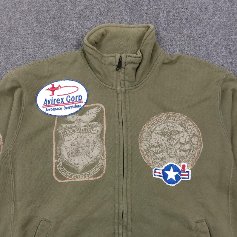 May include: Olive green zip-up jacket with multiple embroidered patches. Patches include the text "Avirex Corp Aerospace Operations", a shield with the text "U.S.S. SARATOGA", and a circular patch with an eagle design. The jacket has a high collar.