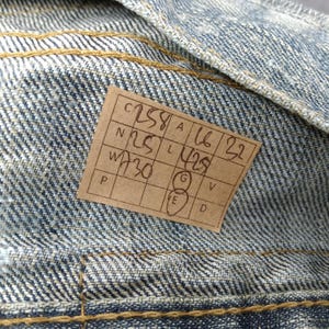 May include: Close-up of denim fabric with visible stitching and a rectangular tag. The tag has handwritten numbers and letters, including "C258", "N25", "W730", "A16", and "L429". The denim is a light blue wash.