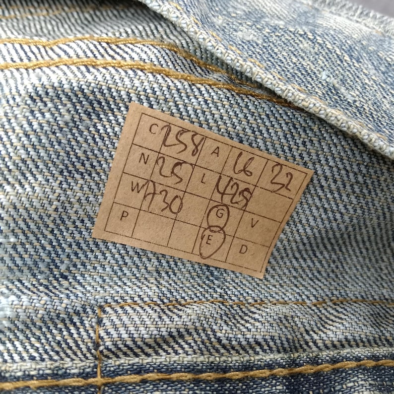 May include: Close-up of denim fabric with visible stitching and a rectangular tag. The tag has handwritten numbers and letters, including "C258", "N25", "W730", "A16", and "L429". The denim is a light blue wash.